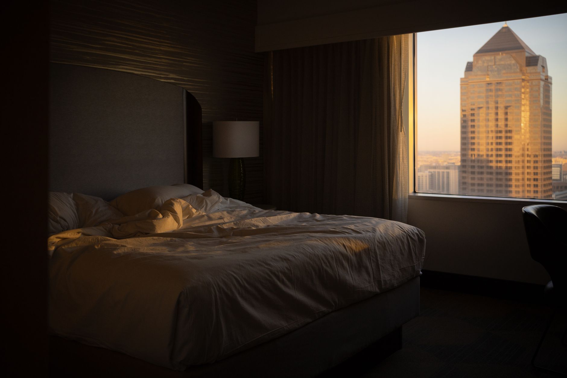An unmade hotel bed in golden evening light beside a window overlooking a city tower