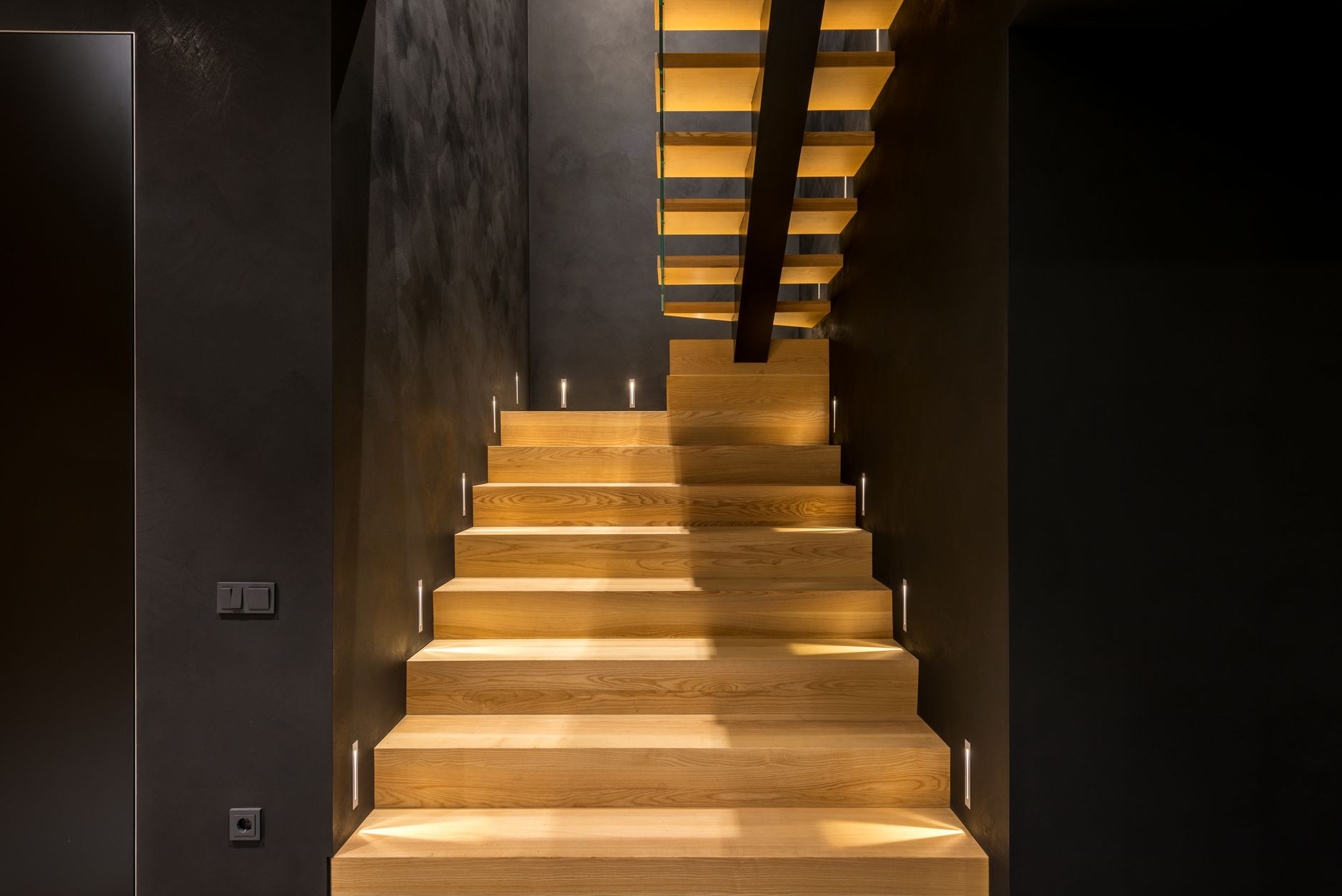A wooden staircase rising in even steps between dark walls, lit by recessed step lights