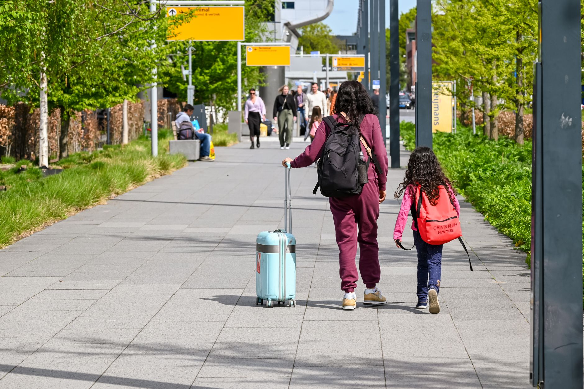 A woman and a child with a suitcase walking along a tree-lined path towards an airport terminal.